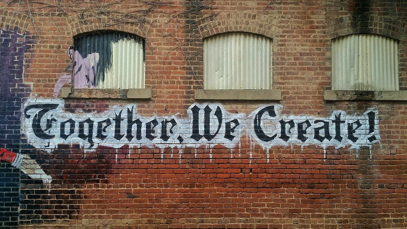 “Together, we create!” graffitied onto a red brick wall.